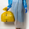 A person wearing a translucent light blue protective gown and blue gloves holds a large yellow biohazard waste bag with black printed labels against a plain light background.